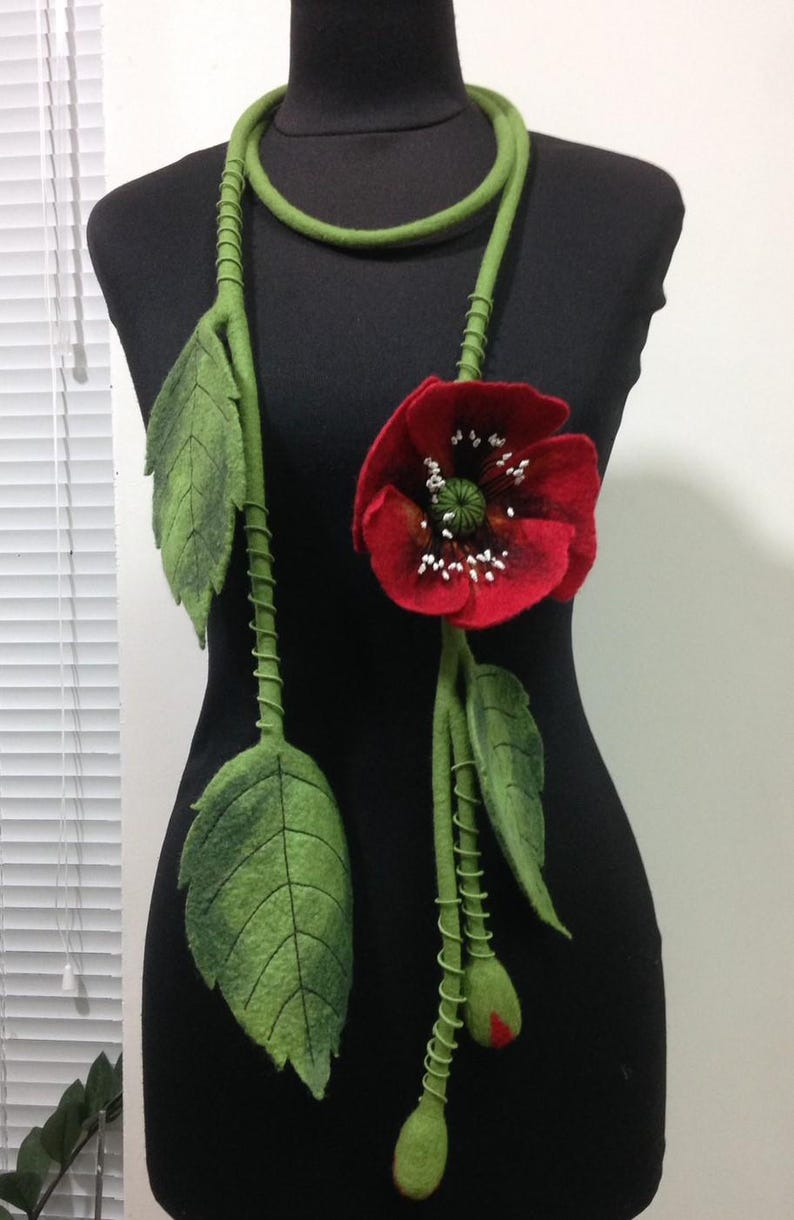 May include: A felt necklace with a red poppy flower and green leaves. The necklace is made of felt and is a unique and stylish accessory.