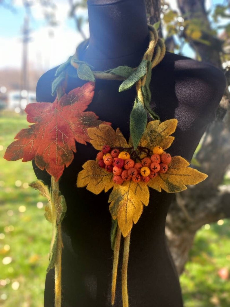 Puede incluir: Un collar de fieltro con una larga hebra de hojas y bayas. Las hojas son verdes, amarillas y naranjas, y las bayas son rojas y naranjas.