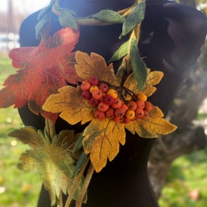 Puede incluir: Un collar de fieltro con una larga hebra de hojas verdes y un racimo de bayas naranjas y rojas. El collar est&aacute; decorado con hojas de fieltro en tonos naranja, amarillo y verde.