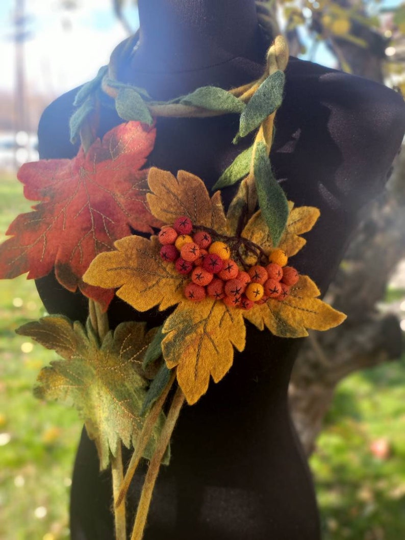 Puede incluir: Un collar de fieltro con una larga hebra de hojas verdes y un racimo de bayas naranjas y rojas. El collar est&aacute; decorado con hojas de fieltro en tonos naranja, amarillo y verde.