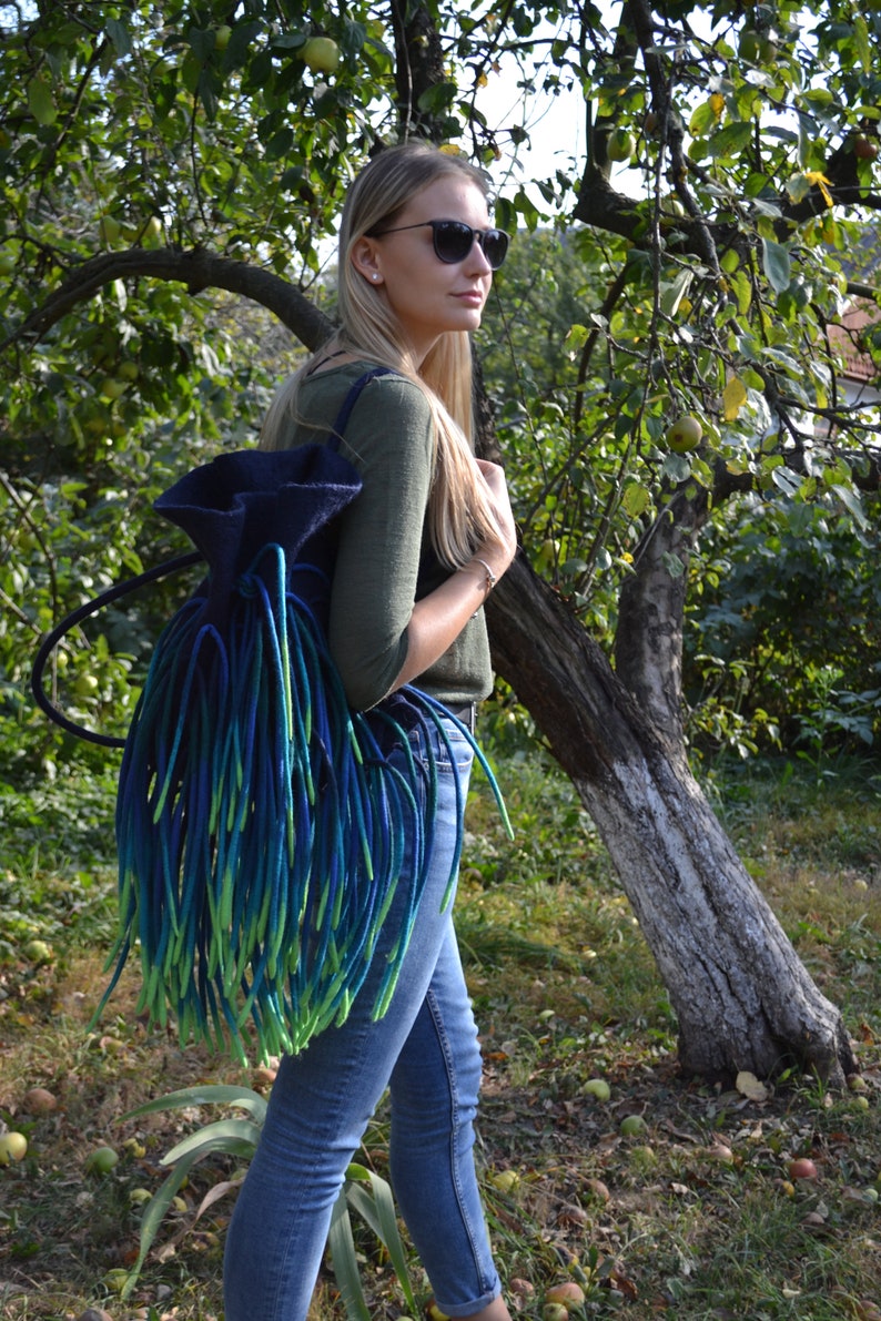 May include: A woman wearing blue jeans and a green long-sleeved shirt is standing in a garden. She is wearing sunglasses and carrying a large, blue and green yarn backpack.