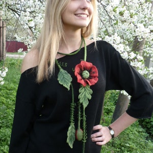 May include: A woman wearing a black sweater with a long, felt flower necklace. The necklace features a red poppy with green leaves and stems.