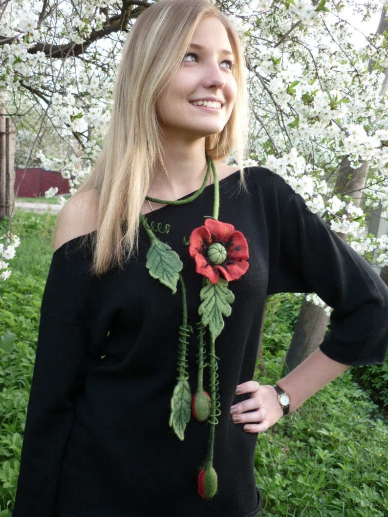 May include: A woman wearing a black sweater with a long, felt flower necklace. The necklace features a red poppy with green leaves and stems.