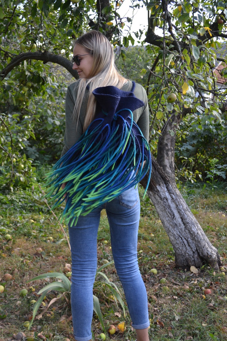 May include: A woman wearing blue jeans and a green shirt is carrying a blue and green felted backpack with long fringe.