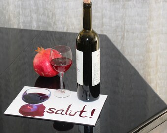 May include: A dark glass wine bottle with a cork stopper, a clear glass filled with red wine, and a whole red pomegranate placed on a rectangular white mat featuring a graphic of a wine glass and the word salut! in red script.