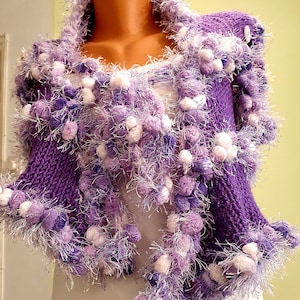 Könnte beinhalten: Ein handgestrickter, violetter Schal mit weißen und lila Pompons und flauschigem Besatz. Der Schal wird auf einer Schaufensterpuppe präsentiert, die sein kompliziertes Design und seine Textur zeigt. Die Farbpalette umfasst Lila- und Weißtöne.