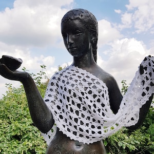 May include: A bronze statue of a woman wearing a white crocheted shawl. The statue is outdoors and the background is a blue sky with white clouds.
