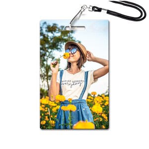 May include: A woman wearing a white t-shirt with the text "Inspire Others Everyday" in black script, blue overalls, a straw hat, and sunglasses, is standing in a field of yellow flowers. She is holding a yellow flower to her nose and smiling.