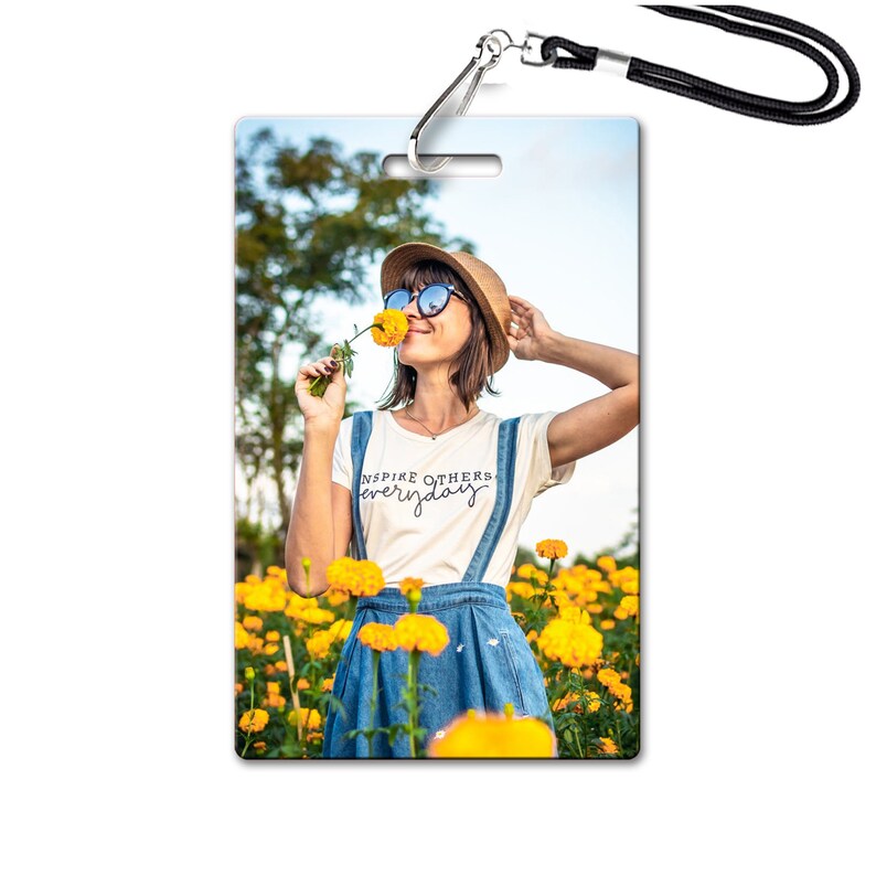 May include: A woman wearing a white t-shirt with the text "Inspire Others Everyday" in black script, blue overalls, a straw hat, and sunglasses, is standing in a field of yellow flowers. She is holding a yellow flower to her nose and smiling.