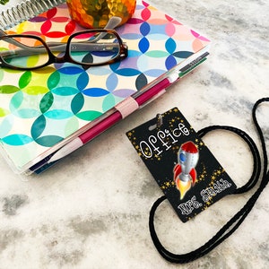 Puede incluir: Un cord&oacute;n negro con un dise&ntilde;o de cohete y estrellas doradas. El cord&oacute;n tiene una etiqueta de nombre en blanco y negro que dice "Office" y "Mrs. Smith".