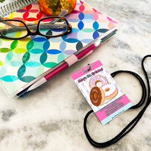 May include: Pink and white birthday party invitation with a black lanyard. The invitation says "You're invited to Abby's 6th Birthday!" and features a graphic of three donuts.