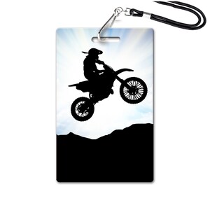 May include: Silhouette of a motocross rider jumping over a hill against a blue sky with clouds. The rider is wearing a helmet and goggles. The motorcycle is black and has a white background.
