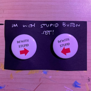 May include: A set of two white buttons with the text "IM WITH STUPID" and a red arrow pointing left and right. The buttons are on a black card with the handwritten text "IM WITH STUPID BUTTON SET!".