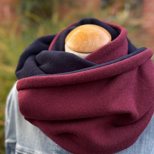 May include: A reversible infinity scarf in burgundy and navy blue. The scarf is made of a soft knit fabric and is draped around a mannequin's neck. The outer layer is burgundy, and the inner layer is navy blue. The scarf is worn over a denim jacket.