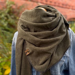 Könnte beinhalten: Ein olivgrüner Schal ist über einer Jeansjacke drapiert. Der Schal besteht aus einem weichen Material und wird mit einem kleinen, quadratischen, braunen Lederknopf befestigt. Der Schal ist um Hals und Schultern gewickelt.