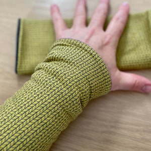 Op de afbeelding: Een hand die een gele gebreide schoonmaakdoek vasthoudt. De doek is getextureerd en heeft een donkergroene rand.