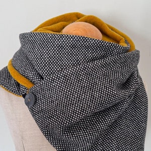 Op de afbeelding: Een zwart-wit gepatroneerde infinity sjaal met een mosterdgele fleece voering. De sjaal heeft een enkele knoopsluiting.