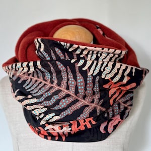Peut inclure: Un tour de cou réversible et douillet avec un extérieur en polaire rouge doux et un intérieur noir orné d'un motif de feuilles de fougère brunes et orange avec des points turquoise.