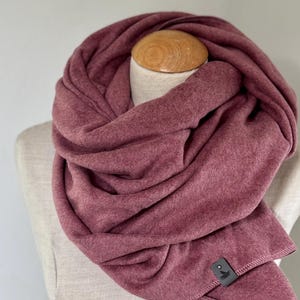 Puede incluir: Bufanda infinita de color rosa polvoriento, exhibida en un maniquí. La suave tela de vellón está envuelta en un bucle continuo. Una pequeña etiqueta gris oscura está cosida en el borde. La bufanda es de color liso y parece cálida y acogedora.