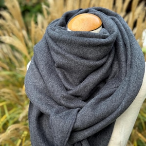 Puede incluir: Bufanda gris oscuro drapeada sobre un maniquí. La bufanda está hecha de un material suave y grueso y está envuelta alrededor del cuello y los hombros del maniquí. El fondo presenta hierbas altas y secas.