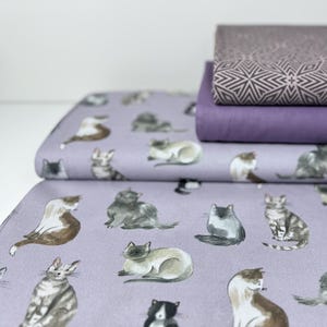 Könnte beinhalten: Lilafarbener Stoff mit einem sich wiederholenden Muster von Katzen in verschiedenen Posen. Die Katzen sind in Aquarelltechnik mit Grautönen, Braun- und Weißtönen gezeichnet. Der Stoff ist dreifach gefaltet, wobei die oberste Schicht ein geometrisches Muster zeigt.