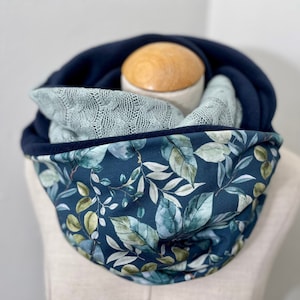 Peut inclure: Un snood bleu marine avec un imprimé de feuilles botaniques dans les tons de vert et de blanc. Le snood est doublé de polaire bleu marine et comporte une écharpe en tricot vert clair à l'intérieur. Un objet en bois est visible à l'intérieur.