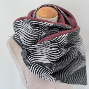 Può includere: Una sciarpa infinita a onde con motivo bianco e nero con bordo bordeaux e un bottone nero.