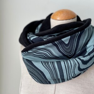 Könnte beinhalten: Ein blauer und schwarzer Infinity-Schal mit einem wellenförmigen, abstrakten Muster. Der Schal ist aus einem weichen, bequemen Stoff gefertigt und hat ein schwarzes Fleece-Futter.