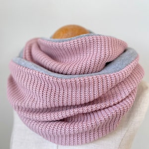 May include: A pink knitted infinity scarf with a grey fleece lining. The scarf is folded and displayed on a white mannequin.