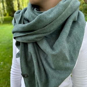 Könnte beinhalten: Ein salbeigrüner Schal, der über einem weißen Langarmshirt drapiert ist. Der Schal besteht aus einem weichen Material und wird mit einem kleinen, dunkel gefärbten Verschluss befestigt. Der Hintergrund ist eine verschwommene Ansicht von grünem Laub.