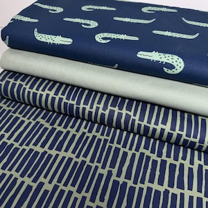 Pode incluir: Uma pilha de rolos de tecido com vários padrões. O rolo superior é azul marinho com um padrão repetido de crocodilos verde claro. Abaixo está um tecido verde claro liso, seguido por um tecido azul marinho com um design de listras verticais verde claro.