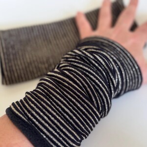 Puede incluir: Una pulsera de mu&ntilde;eca de rayas negras y plateadas, hecha de un tejido suave y el&aacute;stico. La pulsera est&aacute; doblada por la mitad y est&aacute; siendo sostenida por una mano.