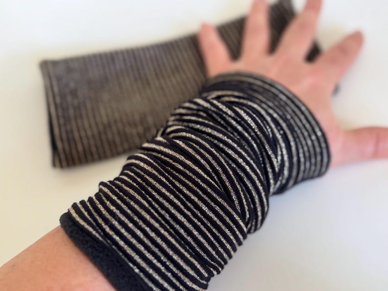 Puede incluir: Una pulsera de mu&ntilde;eca de rayas negras y plateadas, hecha de un tejido suave y el&aacute;stico. La pulsera est&aacute; doblada por la mitad y est&aacute; siendo sostenida por una mano.