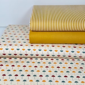 Könnte beinhalten: Drei Stoffmuster, eines mit gelbem und weißem Streifenmuster, eines einfarbig gelb und eines mit weißem Hintergrund und einem sich wiederholenden Muster aus kleinen, bunten Regenschirmen.