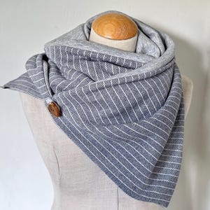 Pode incluir: Cachecol cinza e branco com padrão de espinha de peixe e fecho de botão de madeira. O cachecol é enrolado em volta da cabeça de um manequim e tem um design macio e em camadas. O forro interior é de cor cinza lisa.