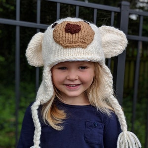 May include: A white knitted bear hat with brown felt ears and nose. The hat has long white braids that tie at the bottom. A young girl is wearing the hat.