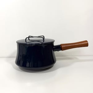 Puede incluir: Un cazo esmaltado de color azul marino oscuro con una tapa a juego que presenta un asa negra en forma de cruz. La olla tiene un acabado liso y brillante, y un mango largo y recto de madera con veteado marrón natural en el lateral.