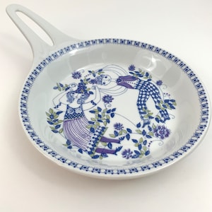 Puede incluir: Un plato de servir de cerámica blanca con un diseño floral azul y morado con tres figuras bailando. El plato tiene un asa y un borde festoneado.