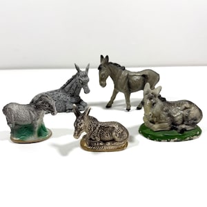 Könnte beinhalten: Fünf Miniatur-Eselfiguren in verschiedenen Posen. Die Esel sind in Grau- und Brauntönen bemalt, einige ruhen auf grünen oder beigen Sockeln. Die Figuren sind wahrscheinlich aus Keramik oder einem ähnlichen Material.