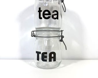 May include: Two clear glass storage jars stacked vertically, each featuring a wire bail closure with a red rubber seal. The top jar has the word tea in black lowercase letters, while the bottom jar displays the word TEA in black uppercase letters.