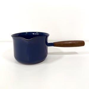 May include: A deep blue enameled metal saucepan featuring a rounded body, a small pouring spout on the rim, and a straight, cylindrical wooden handle attached to the side. The interior and exterior surfaces are finished in a smooth, glossy blue.