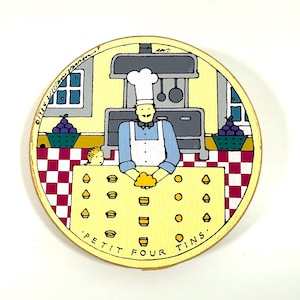 Puede incluir: Una pieza decorativa redonda de madera que presenta una escena de cocina caprichosa. Un chef con gorro y delantal blancos está de pie detrás de una encimera con la inscripción 'PETIT FOUR TINS'. El fondo incluye una estufa, una ventana y cestas de uvas.