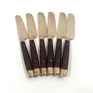 May include: Set of six butter knives. Each knife features a silver blade, a dark brown wooden handle, and a gold-colored metal end. The knives are arranged on a white surface.