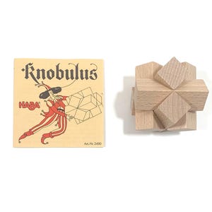 Peut inclure: Un puzzle en bois appelé "Knobulus" de HABA. Le puzzle est un cube à six faces, chacune ayant une forme différente. Le puzzle est en bois brun clair et a la forme d'une étoile.