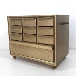 May include: A gold-colored storage cabinet with a textured, glittery finish. The unit features nine small, rectangular drawers arranged in three rows of three, positioned above one large, wide drawer. The cabinet stands on four small, round feet.