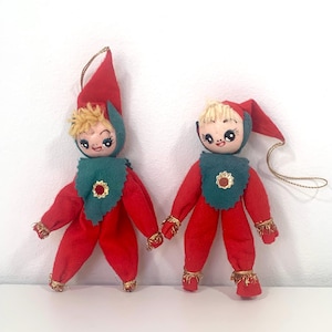 Peut inclure: Deux décorations de Noël en feutre en forme d'elfes. Les elfes portent des costumes rouges avec des accents verts et ont les cheveux blonds. Ils ont de grands yeux et des visages souriants.