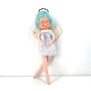 May include: A vintage angel doll with blue hair, closed eyes, and a white dress with a ruffled hem. The doll has fabric wings and a halo. The doll's legs are crossed, and it is standing against a white background.