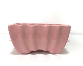 May include: A pink ceramic planter with a wavy, scalloped rim. The planter is made of glazed ceramic and has a smooth, glossy finish.