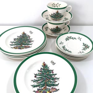 Puede incluir: Un juego de vajilla navideña con un borde verde. El juego incluye platos, platillos y tazas de té, cada uno decorado con una ilustración detallada de un árbol de Navidad. Los platos y tazas son blancos con un borde verde.
