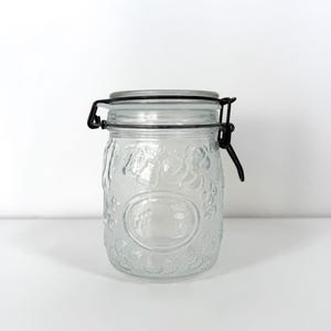 Puede incluir: Un tarro de vidrio transparente con un diseño floral en relieve. El tarro tiene una tapa con bisagras de metal y un cierre de metal negro. El tarro está diseñado para el almacenamiento y tiene una estética vintage.